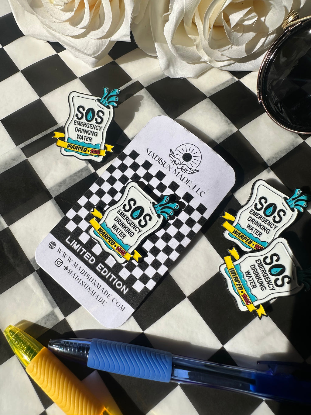 Enamel pin with "SOS Emergency Drinking Water" and yellow banner reading "Warped 2025" on the front with a checkered background