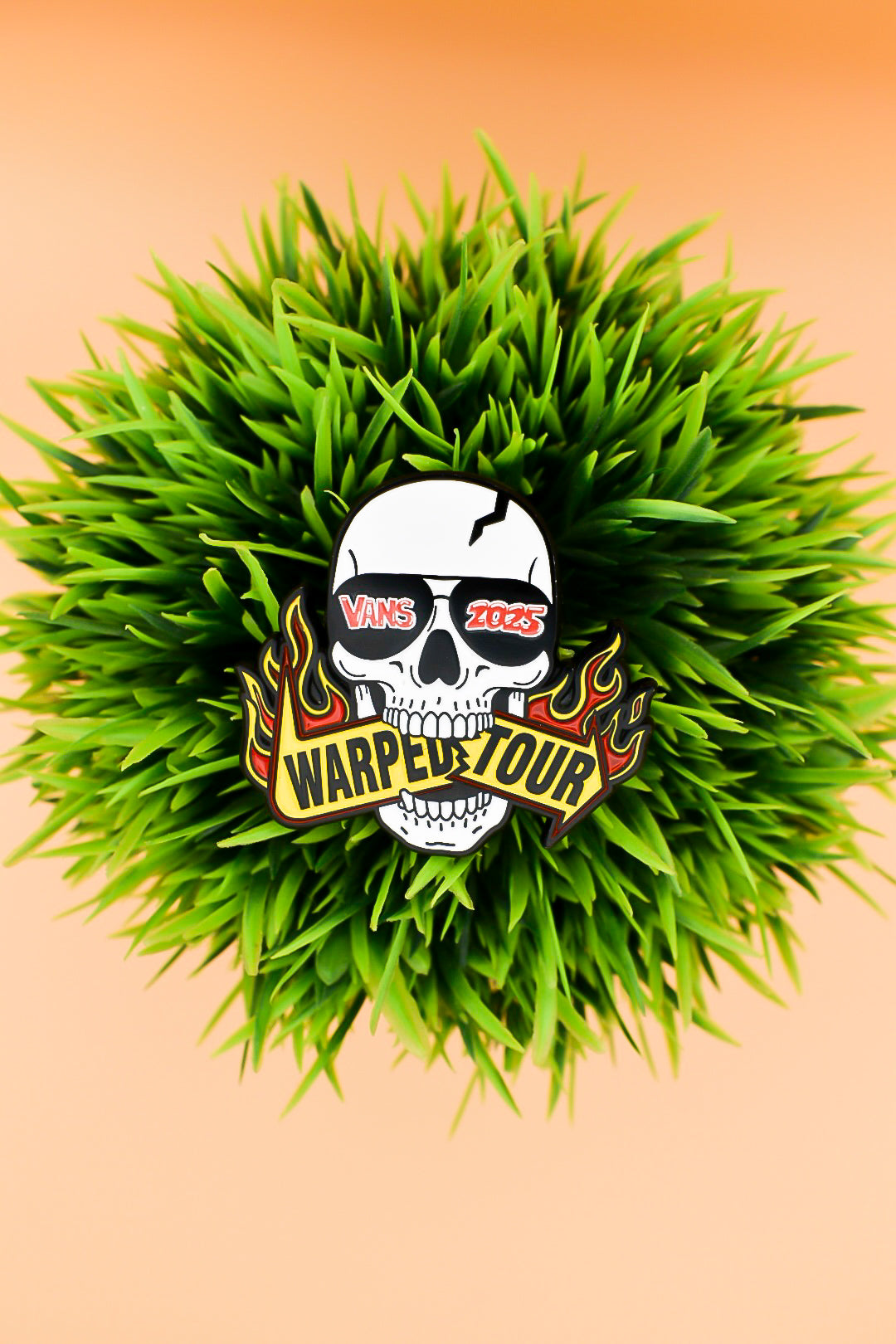 Skull with sunglass that read "Vans 2025" biting a sign that reads "Warped Tour" on grass